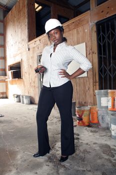 An attractive black woman wearing a white hardhat stands with her cell phone in her hand.