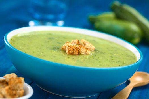 Zucchini cream soup with wholewheat croutons served in blue bowl with wooden spoon on the side, zucchini and glass in the back (Selective Focus, Focus on the front of the croutons) 