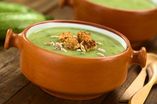 Zucchini cream soup with wholewheat croutons and roasted sunflower seeds served in rustic bowl with wooden spoons on the side (Selective Focus, Focus on the front of the croutons) 