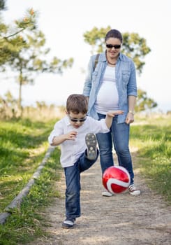 Happy cute son playing with soccer ball in the park, and his pregnacy mother looking in the background