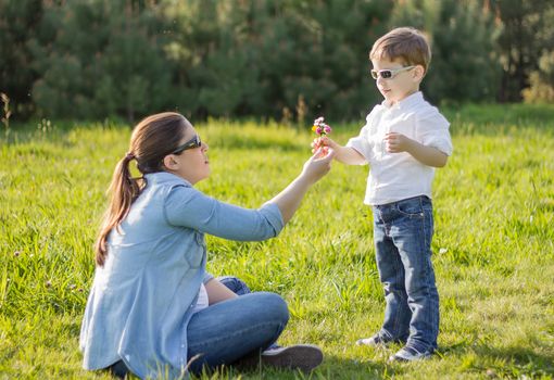 Happy cute son giving a bouquet of flowers to his pregnant mother in a sunny field