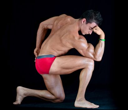 Handsome young man with muscular body in classic pose on his knee, isolated on black