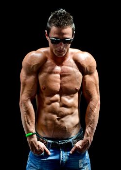Attractive young man with naked muscular torso, wearing jeans and sunglasses, isolated on black background