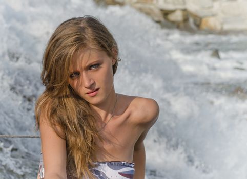 Pretty blonde brunette girl, blue eyes, sitting in front of a river waterfall