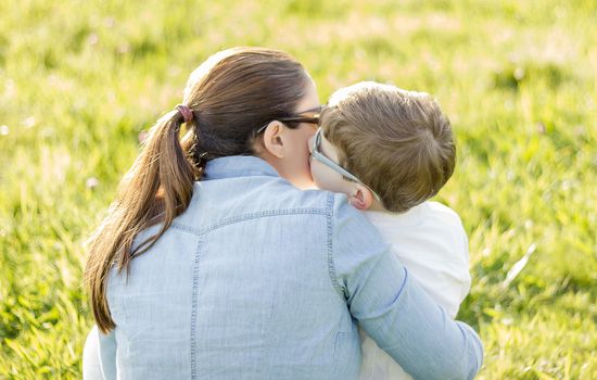 Back view of cute son kissing to his mother sitting in a sunny field
