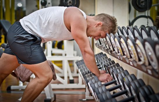 Handsome young athletic man resting on dumbells rack after workout in gym