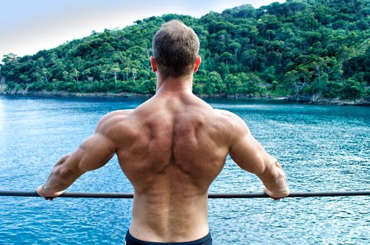 Muscular young man looking at the sea, seen from the back. Leaning on metal railing