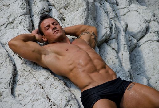 Attractive young man  laying naked on white rocks, eyes closed, wearing only black swimming suit