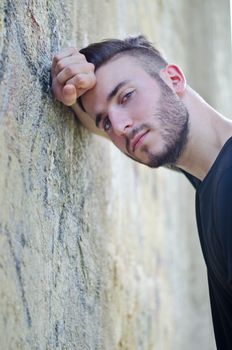 Handsome young man leaning his head against arm on a wall and looking in camera, outdoors