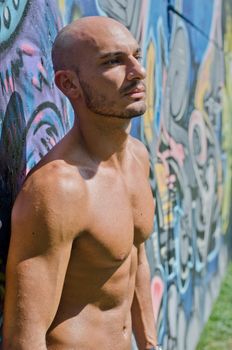 Attractive bald young man shirtless on graffiti wall outdoors in the sun looking to a side