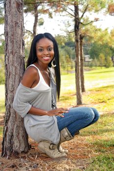 An extraordinarily beautiful young woman with a bright, warm smile leans against a pine tree outdoors.