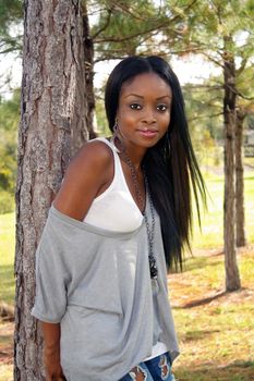 An extraordinarily beautiful young woman with a friendly smile leans against a pine tree outdoors.