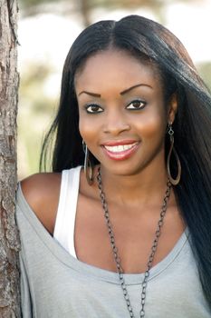 An extraordinarily beautiful young woman with a bright, warm smile leans against a pine tree outdoors.