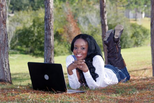 An extraordinarily beautiful young woman with a captivating smile, dressed in casual wear, works on her laptop computer outdoors.  Generous copyspace.