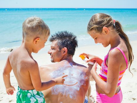 Adorable boy with his sister at tropical beach applying sunblock cream on a father's back.