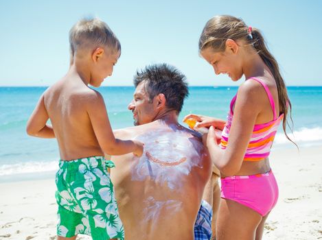 Adorable boy with his sister at tropical beach applying sunblock cream on a father's back.