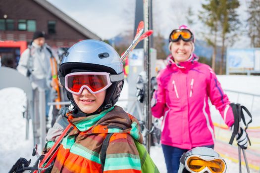 Portrait of happy smiling girl in ski goggles and a helmet with his mother before the ski lift