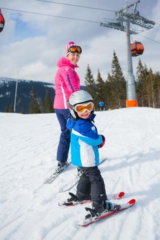 Happy little boy studying to ski in ski goggles and a helmet with his mother