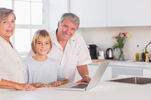 Child and grandparents looking at the camera with a laptop in front in kitchen