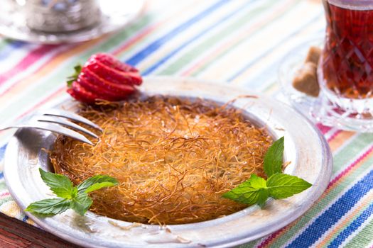 Turkish dessert kunefe  served in classical aluminum plate on a picnic table with mints and sliced strawberry along with Turkish hot tea