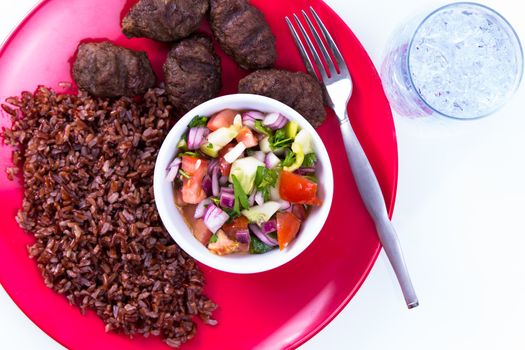 Turkish Meat Balls Kofte served with Red Rice Pilaf and Turkish Shepperd Salad on a red plate along with ice water. Isolated on white