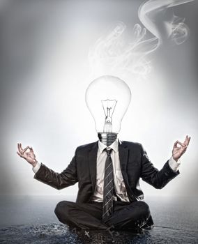 Businessman with bulb head sitting in meditation position with smoke rising on grey background