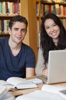 Two students sitting at a desk in a library while using the laptop and smiling