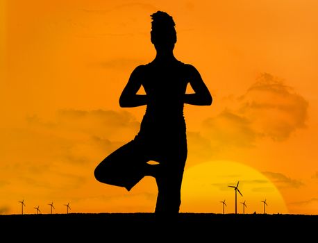 Silhouette of woman doing yoga outdoors in front of sunset and wind turbines