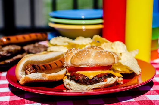 Cookout with cheeseburger, hot dog and potato chips.  Mustard and ketchup bottles, bowls, hamburger patties, and hot dog wieners in background.