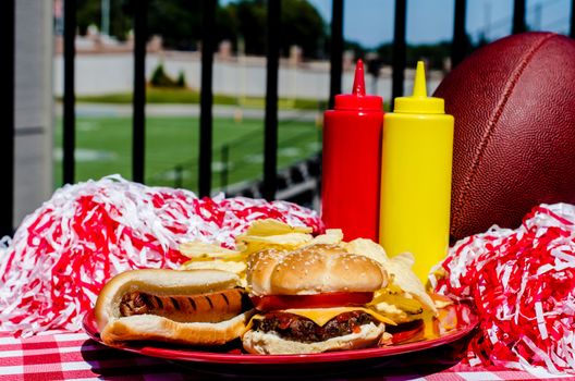 Tailgating party with cheeseburger, hot dog, potato chips, pom poms, and football.  Football field in background.