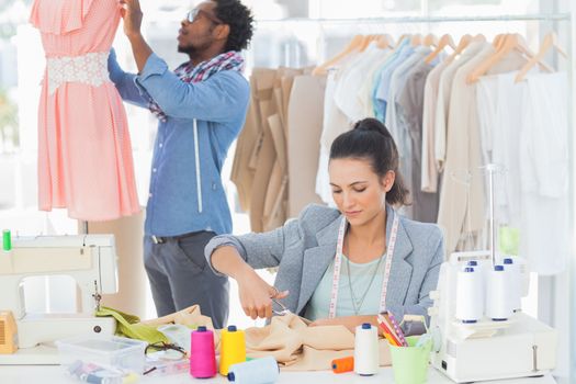 Fashion designer cutting textile at desk while her colleague adjusting dress behind