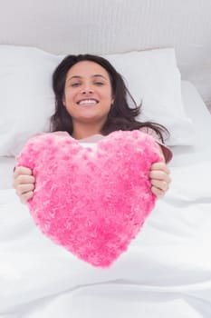 Cheerful woman lying in her bed and holding a fluffy heart cushion