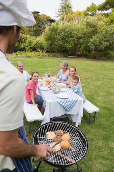 Happy extended family having a barbecue being cooked by father in chefs hat outside in the sun