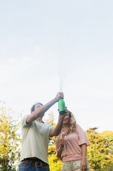 Man spraying bottle of champagne with blonde partner outside in the sunshine
