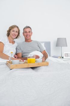 Middle aged couple having breakfast in bed together smiling at camera at home in bedroom