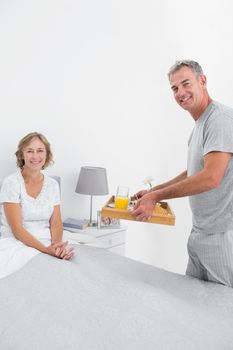 Smiling husband bringing breakfast in bed to wife looking at camera in bedroom at home