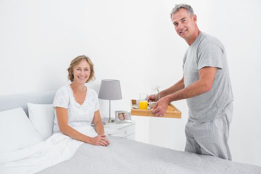Happy husband bringing breakfast in bed to wife smiling at camera in bedroom at home