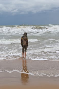 The teenager costs on the seashore in a jacket and barefoot. back in a shot.
