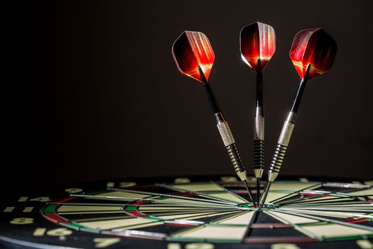 Red, black, and green dartboard on its side with three fiery metal tipped darts in the bullseye. On black background.