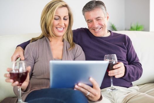 Happy mature couple sitting on a sofa drinking wine and looking at a tablet pc