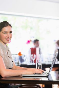 Happy businesswoman typing on laptop and smiling at camera in a cafe