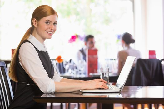 Happy businesswoman typing on laptop while smiling at camera in a restaurant