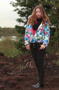 Trendy attractive young woman with long brunette hair standing outdoors in the countryside in a colourful jacket