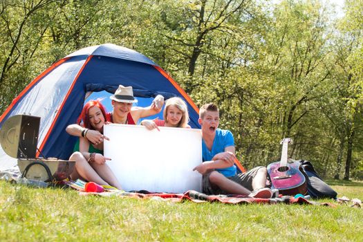 Four teenagers in colored shirts on the camping with a white board which can be used for your advertisements