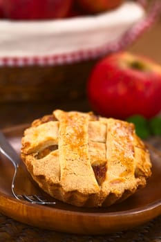 Small round apple pie with lattice crust on wooden plate with pastry fork, in the back an apple and a basket with apples (Selective Focus, Focus one third into the pie) 
