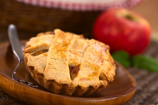 Small round apple pie with lattice crust on wooden plate with pastry fork, in the back an apple and a basket (Selective Focus, Focus one third into the pie) 