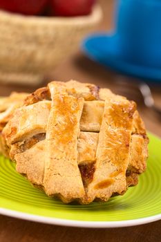 Small round apple pie with lattice crust on green plate, with apples in basket, blue cup and pastry fork in the back  (Selective Focus, Focus one third into the pie) 