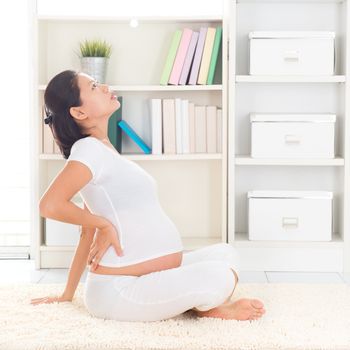 Back pain. Eight months pregnant Asian woman holding her back while sitting on a floor at home.