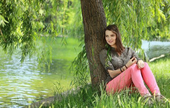 Young woman relaxing under a willow near a pond in a park in summer.