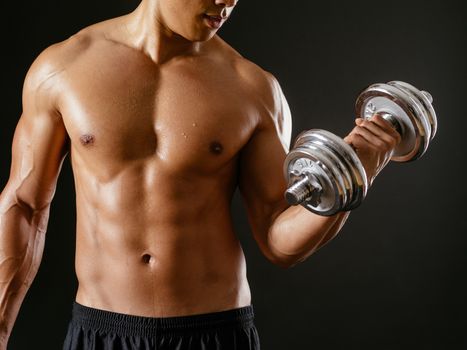 Photo of an Asian male exercising with dumbbells and doing bicep curls over dark background.